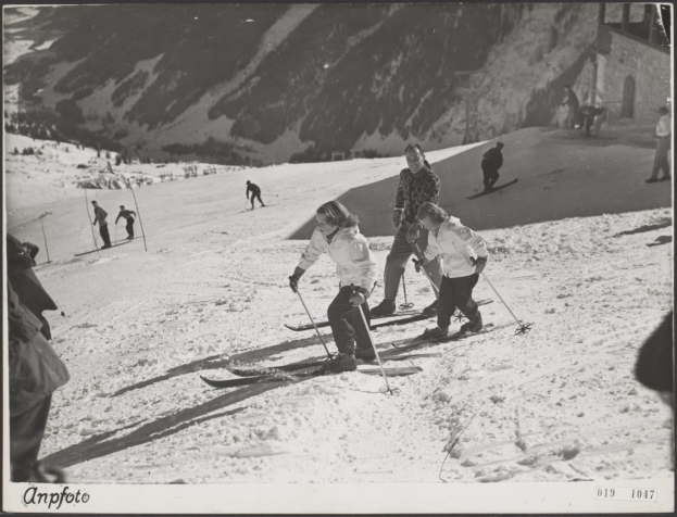 Gruppe von Menschen, die eine schneebedeckte Piste mit Ski-Stöcken hinunterfahren, mit Hügeln und einem Gebäude im Hintergrund und Text am unteren Rand.