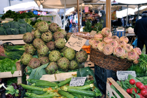 Körbe gefüllt mit Obst, Gemüse und kleinen Schildern mit Text, mit Zelten und wenigen Menschen im Hintergrund.