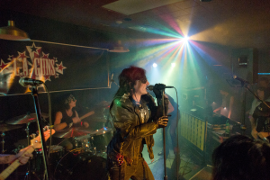 Eine Frau in einer schwarzen Jacke singt in ein Mikrofon auf der Bühne, mit einem Mann in Sonnenbrille und einem Stirnband, der ein Instrument spielt, und einem Plakat mit 'LA Guns' im Hintergrund, unter Diskoleuchten und hängenden Lichtern.