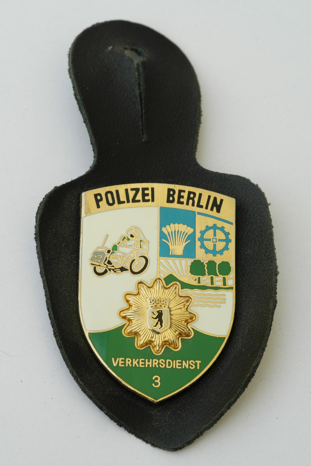 Ein schildförmiges Polizeibadgebnis mit der Aufschrift "Polizei Berlin" auf einem blauen Hintergrund, einem weißen Rand, einem zentralen weißen Stern und schwarzer fetter Schrift, das auf einer Oberfläche liegt.