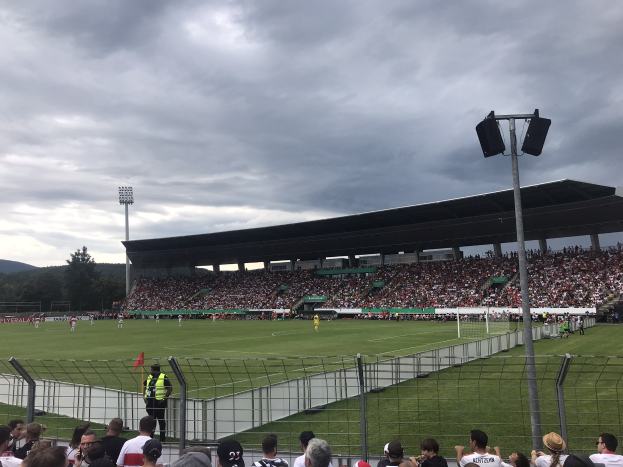 Große Zuschauermenge bei einem Fußballspiel in einem bewölkten, beleuchteten Stadion umgeben von Bäumen und Hügeln.