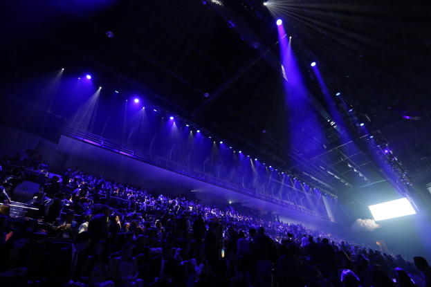 Eine große Menschenmenge in einem Konzertsaal, der einer Bühne mit Deckenleuchten und einem Bildschirm auf der rechten Seite zugewandt ist.