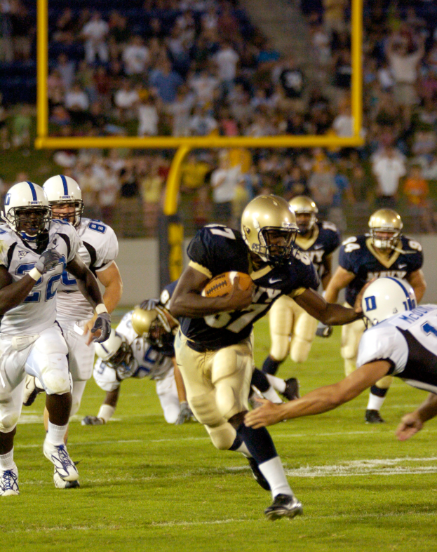 Ein Fußballspieler in einem Sportdress und Helm läuft mit dem Ball, während er von anderen Spielern umgeben ist, mit unscharfen Hintergrundelementen und Text am unteren Rand.