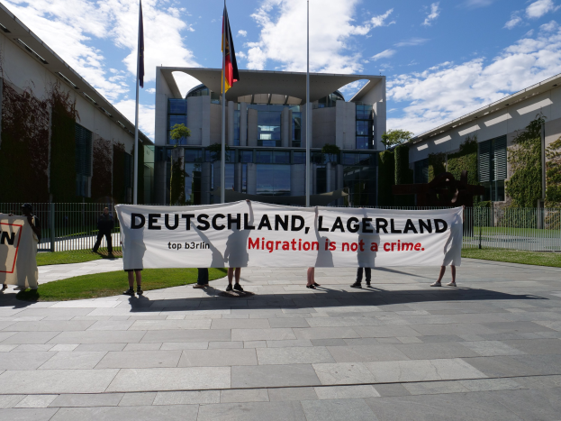 Eine Gruppe von Menschen hält ein Transparent mit der Aufschrift "Deutschland, Lagerland Migration ist kein Verbrechen" vor einem Gebäude mit Fenstern, umgeben von Gras, einem Metallzaun, Pflanzen, Bäumen und Fahnen mit Stäben, unter einem bewölktem Himmel.