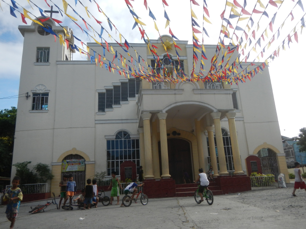 Gruppe von Menschen, die Fahrräder vor einer weißen Kirche mit architektonischen Details, Pflanzen, einem Zaun, einer Fahne, Bäumen, einem bewölkten Himmel und bunten Fahnen an einem Seil über fahren.