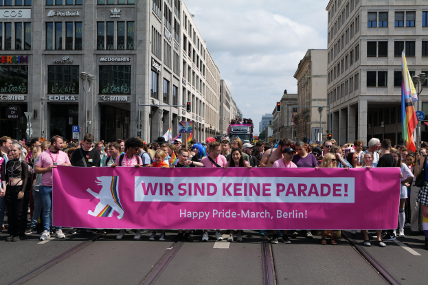 Eine Gruppe von Menschen marschiert auf einer Straße in Berlin, Deutschland, mit einem pinken "Happy Pride March"-Schild, während Gebäude, Laternenpfähle und Verkehrszeichen die Straße säumen und der Himmel bewölkt ist.