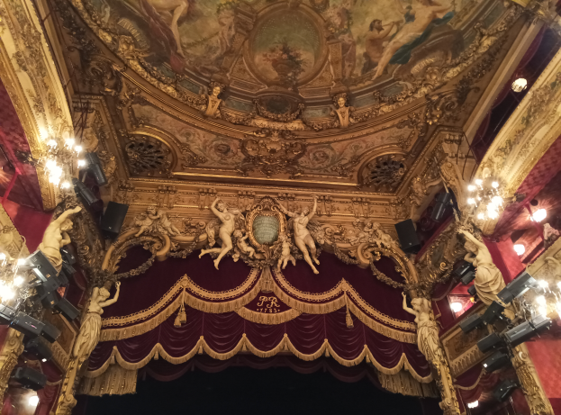 Theaterinnenraum mit roten Vorhängen, Kronleuchtern, Deckenmalerei und Skulpturen, mit einer Bühne und einigen Menschen davor.