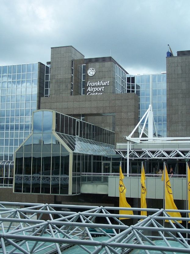 Großes Glasgebäude des Frankfurter Flughafens mit Text darauf, umgeben von gelben Flaggen und Eisenstangen, unter einem teilweise bewölkten Himmel.