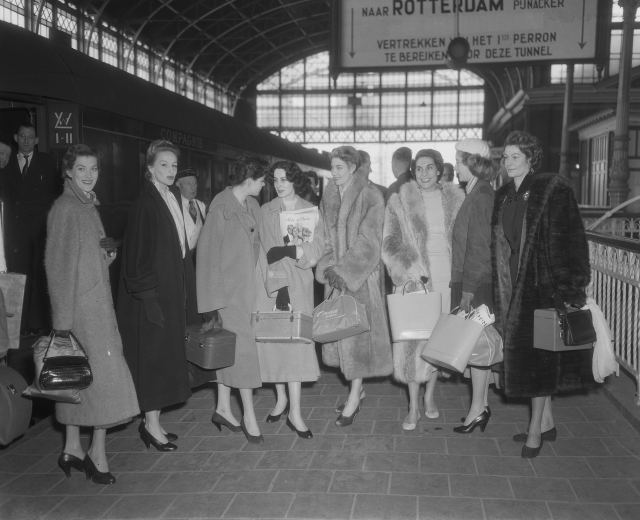 Schwarzes und weißes Bild von Frauen, die an einem Bahnhof mit Taschen stehen, ein Zug auf der linken Seite und Geländer mit einer Tafel auf der rechten Seite.