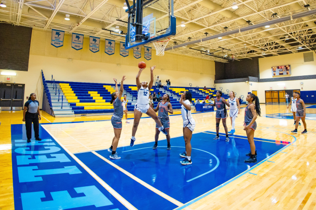 Frauen beim Basketballspielen in einer Turnhalle mit Zuschauern, einer Anzeigetafel mit einem Turniersieg und Bannern an der Wand.