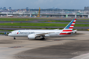 Ein Flugzeug der American Airlines steht auf der Rollbahn eines Flughafens, umgeben von Gras, Fahrzeugen, Gebäuden, Polen, Kränen und anderen Gegenständen, mit Bäumen und einem klaren blauen Himmel im Hintergrund.