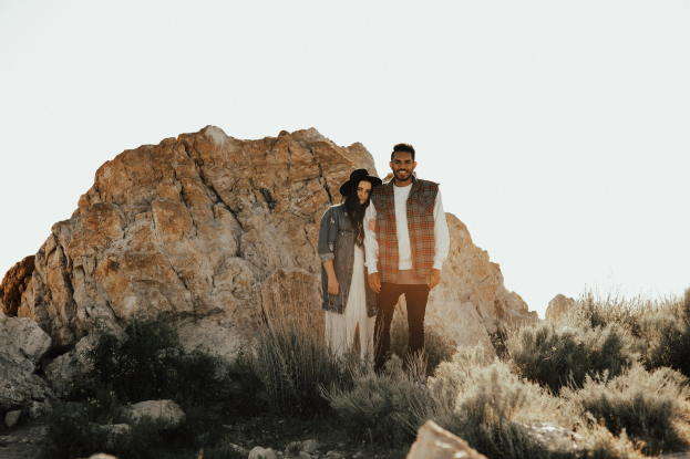 Ein Mann und eine Frau, die auf einem Felsen stehen und in die Kamera lächeln, umgeben von Wüstenvegetation unter einem klaren blauen Himmel während einer Verlobungsfotoshoot.