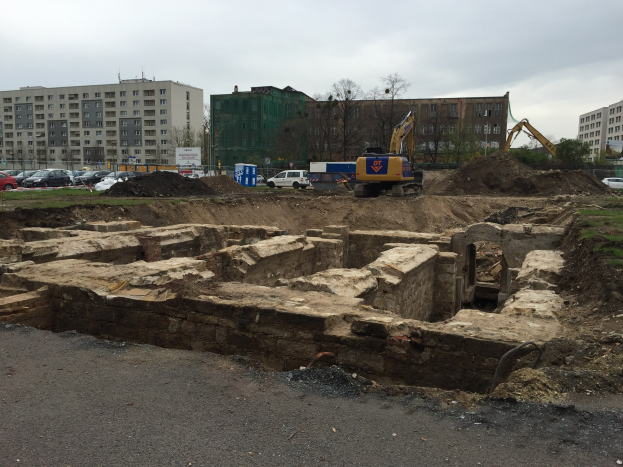 Eine Baustelle mit einem großen Haufen Erde, Baggern, Fahrzeugen, Gras, Bäumen, Gebäuden mit Fenstern im Hintergrund, einem bewölkten Himmel und einer Straße unten, wahrscheinlich Teil einer Grabung in Berlin.