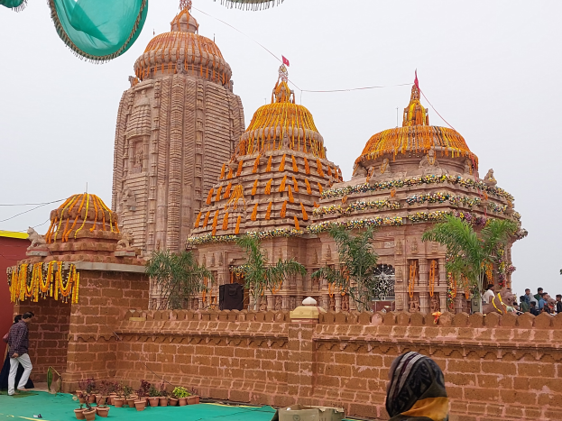 Eine Gruppe von Menschen vor einem Hindu-Tempel mit bunten Girlanden und Flaggen, umgeben von üppiger Vegetation und einem strahlend blauen Himmel, die sich um einen grünen Teppich versammeln.