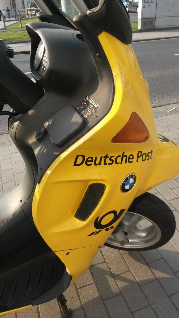 Ein gelber Scooter steht am Straßenrand mit einem sichtbaren Kfz-Kennzeichen, vor Häusern, Bäumen, Pfählen, Schildern und anderen Fahrzeugen auf der Straße.