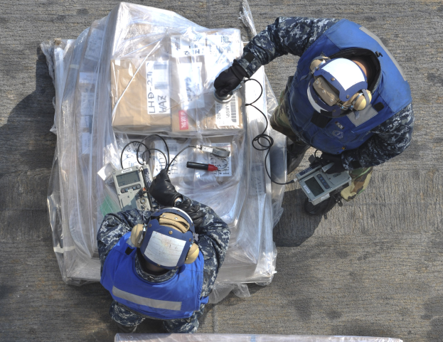 Zwei Männer in blauen Uniformen und Helmen arbeiten an einem großen Drohnen, halten verschiedene Gegenstände, mit einer Plastikabdeckung auf dem Boden.