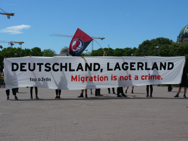 Eine Gruppe von Menschen, die draußen mit einem Banner stehen, auf dem "Deutschland, Lagerland Migration ist kein Verbrechen" steht, und eine Flagge, mit Straßenlaternen, Bäumen, Kränen, einem Gebäude und einem bewölkten Himmel im Hintergrund.