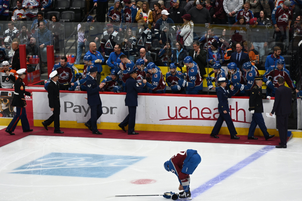 Eishockeyspieler auf dem Eis während eines Spiels zwischen Colorado Avalanche und San Jose Sharks, umgeben von Teamkollegen und Zuschauern, mit einem Scoreboard im Hintergrund, das das Datum und die Begegnung anzeigt.