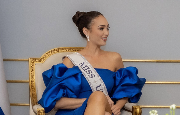 Frau in einem blauen Kleid mit einem "Miss Universe 2019"-Band, die lächelt, während sie auf einem Stuhl sitzt und von Blumen umgeben ist, mit einer Wand im Hintergrund.