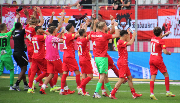 Gruppe von Fußballspielern, die ein Tor feiern, auf einem Feld, einige halten Flaschen oder klatschen, mit Bannern, einem Zaun, Metallstangen, Stühlen und einer Decke mit Deckenleuchten im Hintergrund.