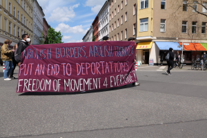 Eine Gruppe von Menschen marschiert mit einem Banner, auf dem "Abolish Borders, Abolish Frontiers, Put an End to Deportations, Freedom of Movement 4 Everyone" steht, durch eine Straße mit Gebäuden, Bäumen, Fahrrädern und bewölktem Himmel im Hintergrund.