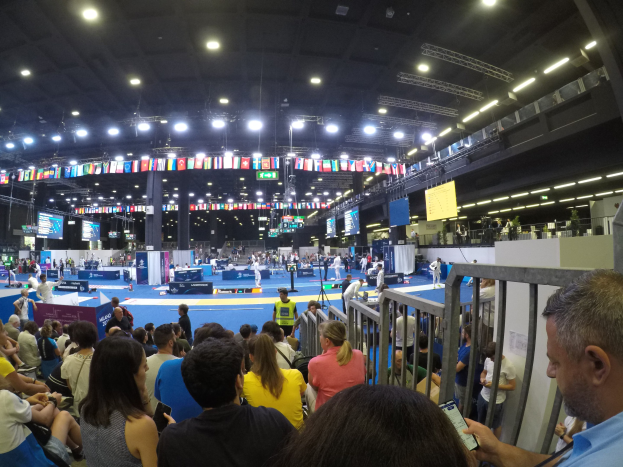 Eine große Menschenmenge auf Sitzplätzen und Stehplätzen in einer Indoor-Sportarena mit Bildschirmen, Flaggen und Deckenleuchten im Hintergrund.
