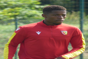 Ein junger Mann in einem roten und gelben Trikot steht auf einem Fussballfeld umgeben von einem Zaun und Bäumen und sieht entschlossen aus, als wäre er ein Flügelspieler.