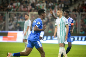 Ein Fußballspieler in einem blauen und weißen Trikot feiert ein Tor, umgeben von einer Gruppe in weißen und grünen Trikots, mit einem unscharfen Hintergrund, der ein weißes und blaues Brett und ein Geländer zeigt.