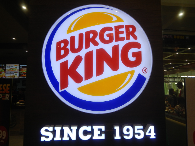 Ein Burger King-Schild in einem Speiseraum mit Menschen und anderen Gegenständen sowie einer Speisekarte mit dem Burger King-Logo links daneben.