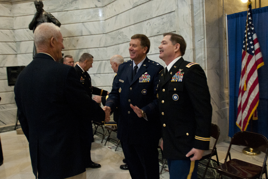 Menschen schütteln sich die Hände vor Stühlen, mit einer Statue, Flagge und Vorhang im Hintergrund.