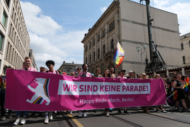 Eine Gruppe von Menschen marschiert auf einer Straße in Berlin, Deutschland, mit einer pinken Fahne mit der Aufschrift "Happy Pride March", mit Gebäuden auf beiden Seiten und einem Fahnenmast im Vordergrund unter einem bewölkten Himmel.