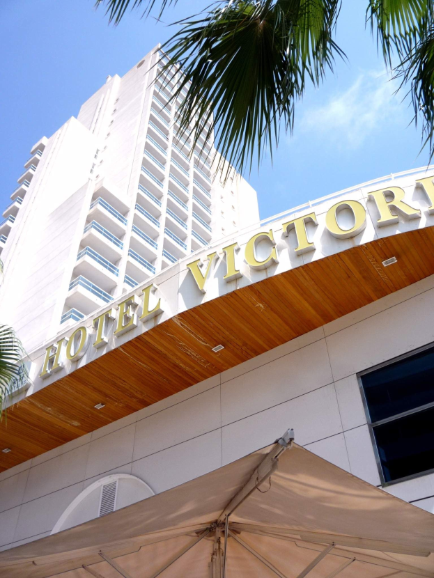 Das Bild zeigt das Hotel Victoria in Miami Beach, Florida, mit einem hohen Gebäude mit Glasfenstern und einer Namenstafel an der Wand, einem Zelt und einem Baum unten und einem Himmel voller weißer Wolken im Hintergrund.