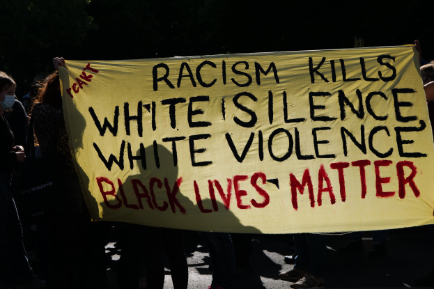 Eine Gruppe maskierter Individuals auf einer Straße bei Nacht, die eine Fahne halten, auf der steht: "Rassismus tötet Weiße Stille, weiße Gewalt, Black Lives Matter."