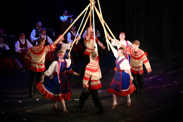 Eine Gruppe von Menschen in farbenfrohen Kostümen tanzt um einen Maibaum, einige halten hölzerne Stöcke, während Musiker im Hintergrund Instrumente spielen.