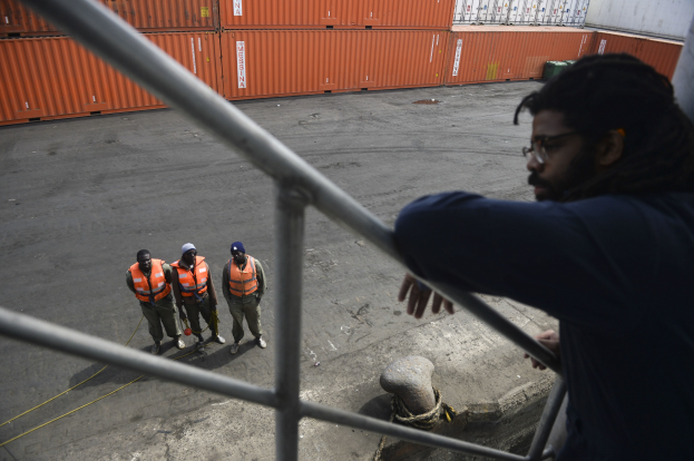 Ein Mann in legerer Kleidung steht auf einem Metallgeländer neben einer Gruppe von Menschen in orangefarbenen Jacken und Mützen, mit Containern und einer Wand im Hintergrund am Hafen von Los Angeles.