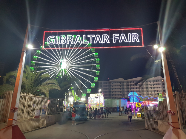 Der Eingang zur Gibraltar-Messe bei Nacht, mit einem hell erleuchteten Riesenrad und einer leuchtenden Namensanzeige, umgeben von Menschenmassen, Bäumen und Gebäuden im Hintergrund unter einem dunklen Himmel.