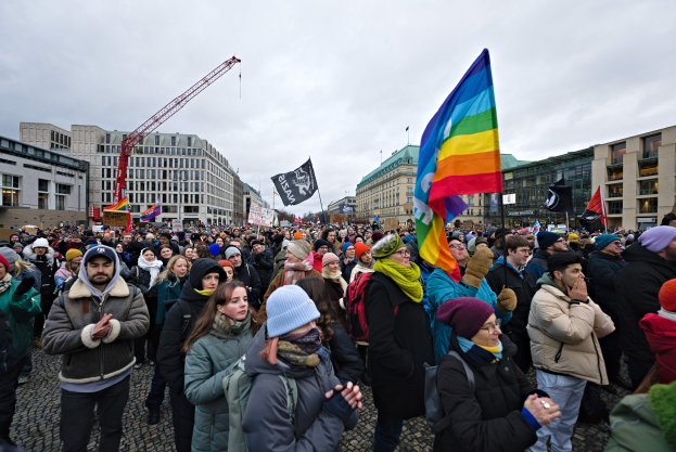 Große Gruppe von Menschen mit Fahnen und Schildern mit Text, einschließlich "Lgbtq+ Rechte Demonstration in Berlin", vor einem Gebäude mit Fenstern, einem Kran und einem bewölkten Himmel.
