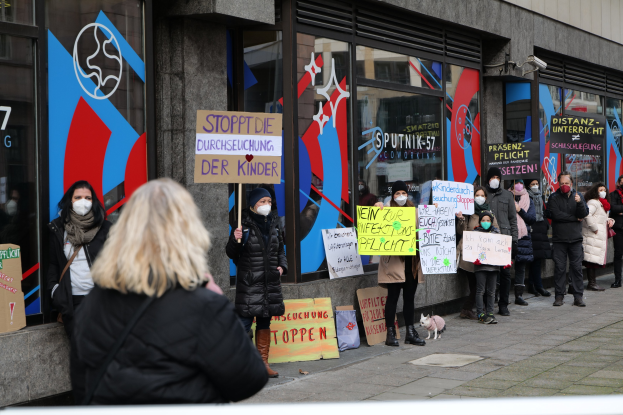 Eine Gruppe von Menschen mit Masken und Schildern protestiert vor einem Glasgebäude, mit Texttafeln, Kameras und einem Hund.