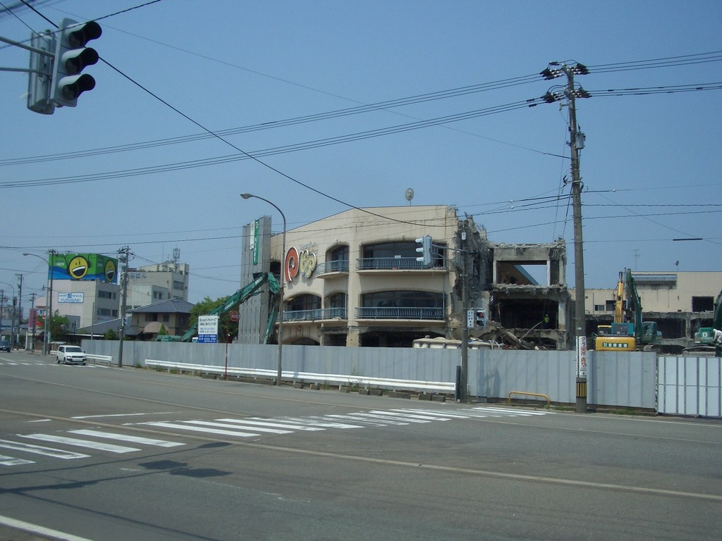 Stadtstraße mit fahrenden Fahrzeugen, Strommasten mit Drähten, Straßenlaternen und einer Ampel auf der linken Seite, flankiert von Gebäuden, Bäumen und Himmel, mit einem teilweise abgerissenen Gebäude im Vordergrund.