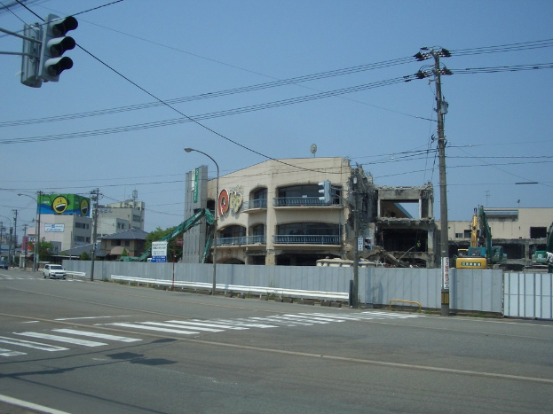 Stadtstraße mit fahrenden Fahrzeugen, Strommasten mit Drähten, Straßenlaternen und einer Ampel auf der linken Seite, flankiert von Gebäuden, Bäumen und Himmel, mit einem teilweise abgerissenen Gebäude im Vordergrund.