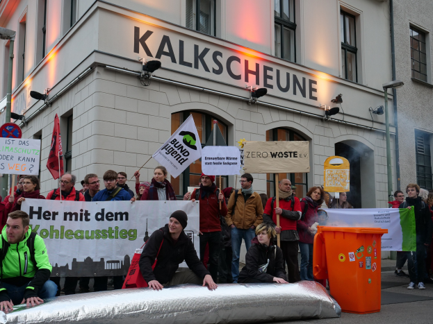 Eine Gruppe von Menschen demonstriert vor einem Gebäude in Deutschland, mit Schildern und Plakaten, mit zwei Personen im Vordergrund und einem Müllcontainer rechts.