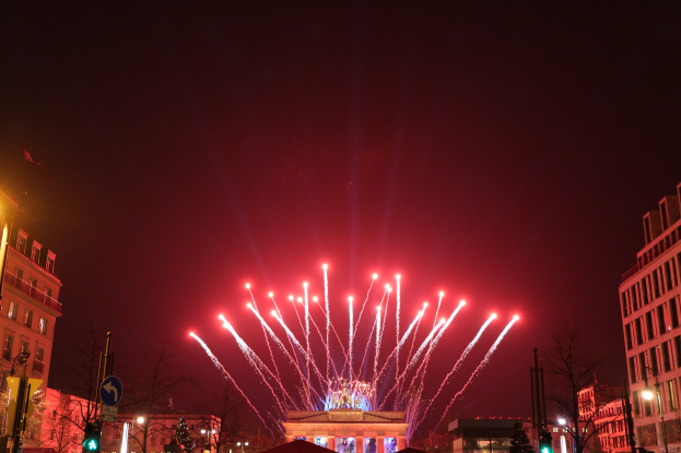 Eine nächtliche Stadtstraße in Berlin, Deutschland, mit dem Brandenburger Tor in der Mitte, umgeben von beleuchteten Gebäuden, Bäumen, Polen, Verkehrszeichen, Schildern, Flaggen und Feuerwerk, das den Himmel erhellt.