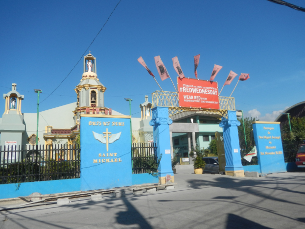 Kircheneingang mit einem Turmuhr im Hintergrund, umgeben von einem Metallzaun, Topfpflanzen, Fahrzeugen, einem Schild, Pfosten, Drähte, einem Straßenpfosten, Bäumen und einem bewölkten Himmel.