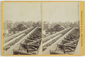 Schwarz-weiß-Foto einer Zugstrecke mit Gebäuden im Hintergrund, Text 'Merkelbach & Co, Co. Amsterdam' rechts und verstreute hölzerne Gegenstände im Vordergrund.
