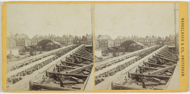 Schwarz-weiß-Foto einer Zugstrecke mit Gebäuden im Hintergrund, Text 'Merkelbach & Co, Co. Amsterdam' rechts und verstreute hölzerne Gegenstände im Vordergrund.