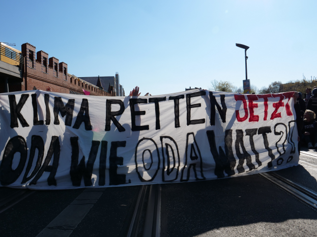 Eine Gruppe von Menschen steht auf der Straße und hält ein Banner mit der Aufschrift "Klima Retten Jetz Oda Wie Oda Watt?" mit Gebäuden, Bäumen und einem Laternenpfahl im Hintergrund.