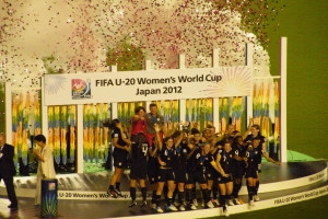 Gruppe von Frauenfußballspielerinnen in schwarzer Uniform auf einer Bühne feiernd, umgeben von einer Menge mit Kameras, mit einem dekorierten Hintergrund und grasbewachsenem Boden.
