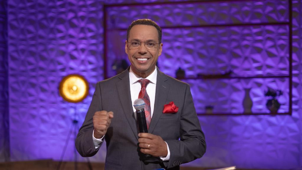 Stephen Colbert, Moderator von "The Daily Show with Trevor Noah", steht auf einer Bühne und hält ein Mikrofon, trägt einen Anzug, eine Krawatte und eine Brille, während im Hintergrund ein Scheinwerfer und ein Ständer zu sehen sind.