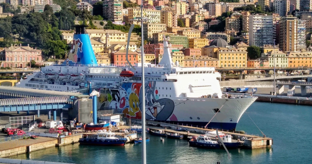 Ein großes Kreuzfahrtschiff liegt nahe einer Stadt, mit einer Brücke links, Fahrzeugen auf der Straße und Gebäuden und Bäumen im Hintergrund.