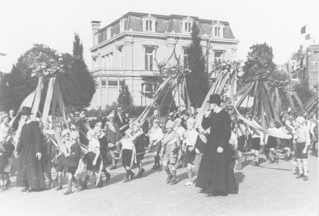 Schwarzes und weißes Bild einer Gruppe von Menschen, die über eine Straße gehen, einige halten Fahnen, mit Hüten, Bäumen, Gebäuden und einem Maibaum im Hintergrund.